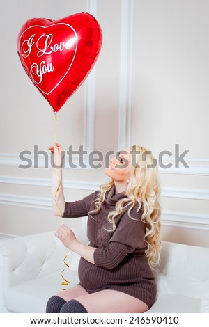 young beautiful pregnant woman sitting on a sofa and looking to the balloon in the form of heart in hand. Valentine\'s Day.