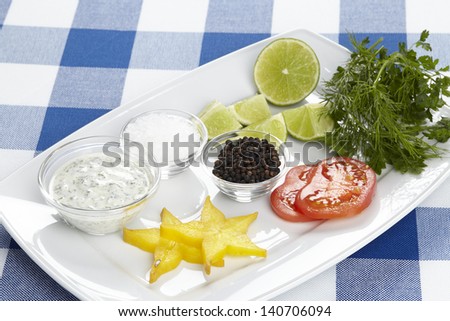 A plate with starfruit slices, tomato slices, dip, salt, black pepper, lime pieces and slices, dill and parsley.