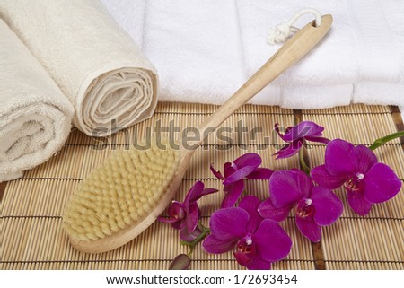 A bath brush is laying on a pile of white, folded towels. Beneath are two naturally colored, rolled towels and a purple orchid.