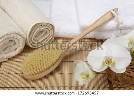 A bath brush is laying on a pile of white, folded towels. Beneath are two naturally colored, rolled towels and a basket with a white orchid.