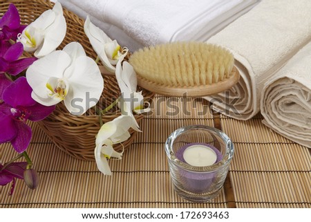 A bath brush with purple and white orchids into a basket standing in front of some rolled towels. The scene is decorated with a tealight into a glass bowl. All objects are standing on a mat of bamboo.