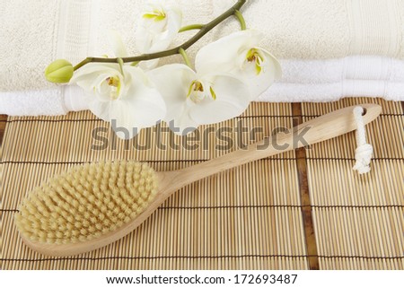 A bath brush is laying on a mat of bamboo. In the background are two pile of folded towels decorated with a white orchid.