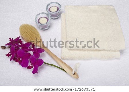 A bath brush is laying beneath two folded, naturally colored towels. The decoration is made of a purple orchid and two tealights into a glass bowl.