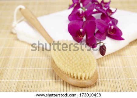 A bath brush is laying on a white folded towel. The scene is taken on a bamboo mat and decorated with a purple orchid.