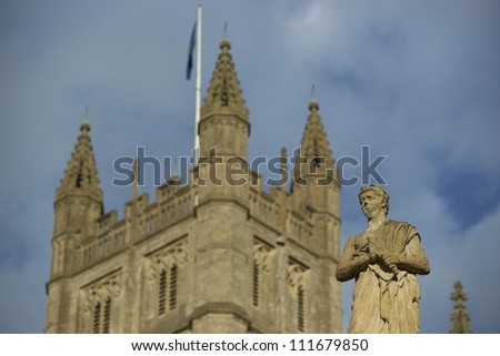 Tower of Bath Abbey standing tall above a weathered stone statue of the ancient Roman Baths in Bath, Somerset, England.