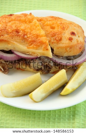 Steak Sandwich Cheese steak sandwich on a round white plate.  The sandwich is on a cheese bun, with red onion.  There are three pickle slices next to the sandwich.