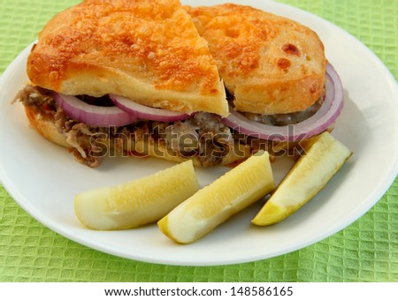 Steak Sandwich Cheese steak sandwich on a round white plate.  The sandwich is on a cheese bun, with red onion.  There are three pickle slices nest to the sandwich.