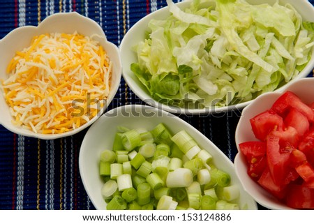 Cheese, Scallions,Lettuce and Tomato Three white serving bowls with shredded cheese, sliced scallions, shredded lettuce and sliced tomato.  The background of the image is textured striped dark blue.
