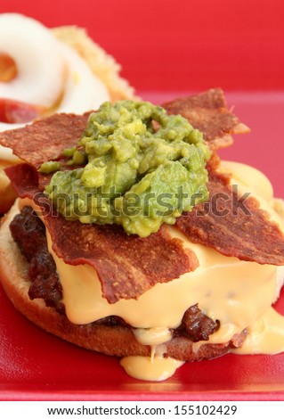 Bacon Cheeseburger and Guacamole Delicious burger with american cheese,bacon,and guacamole on top.  The cheeseburger is on red plate.  The background is the bun top with ketchup and onion slices.