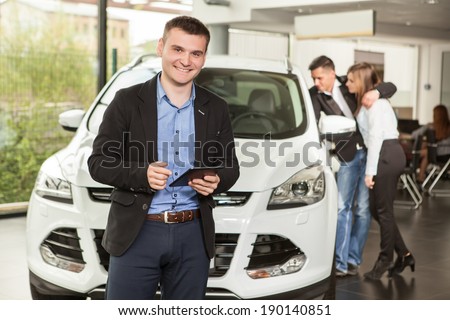 Let me assist you in your vehicle search. Handsome young classic  salesman standing at the dealership with the customers on the background