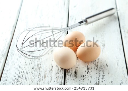 Stainless steel egg whisk with eggs on white wooden background