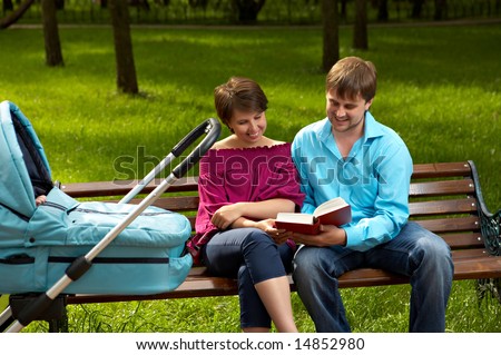 Married couple on walk with the baby sit on a bench in park and read the book on a background of a lawn