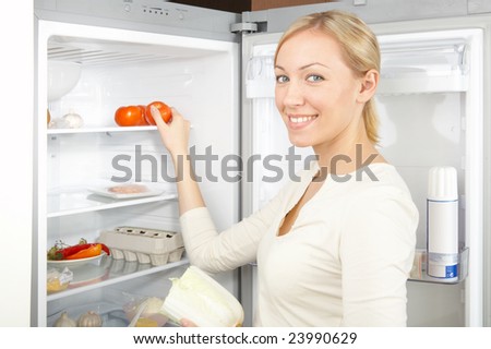 A smiling blonde takes a tomato from a refrigerator