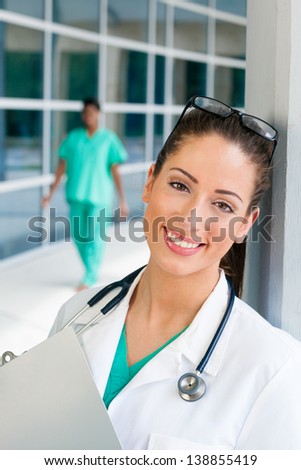 Smiling Caucasian doctor holding a clipboard in brightly lit exterior hospital environment in scrubs, white lab coat and holding glasses. Nurse in scrubs in the background.