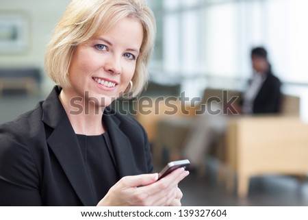 Blonde businesswoman wearing suit in lobby, holding cell phone, looking at camera, smiling