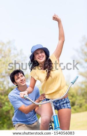 Young latin man in shorts and blue shirt helps young woman riding blue bike with green trees and grass. Horizontal shallow focused composition.