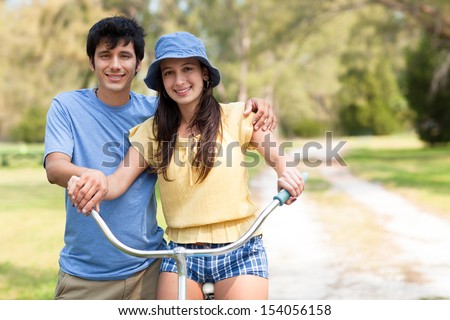Young latin man in shorts and blue shirt posing with young woman riding blue bike with green trees and grass. Trial on background. Horizontal shallow focused composition.