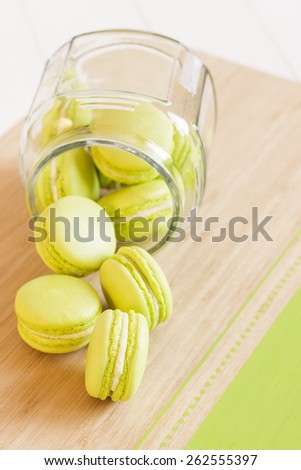 Green macaroons spill out of a glass jar laying on its side. Objects located on the left side pictures are diagonally. Selective focus on first macaroon