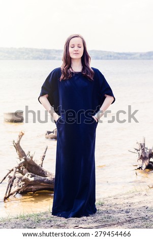 Beautiful young model in long navy blue dress posing at summer beach near water