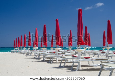 Many folded red umbrellas are aligned on a white sand beach, under a deep blue sky.