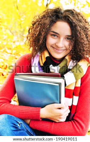 A smiling cute young female student with books in hand on the background of the autumn landscape. Female student in warm autumn.