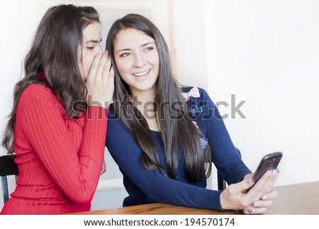 Two young woman discuss something. One from them whispers  something to other girl, who holds in her hand phone.