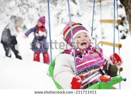 Happy children playing in the winter. Little girl sitting on a swing in a winter garden and children are playing in the background. It\'s snowing.