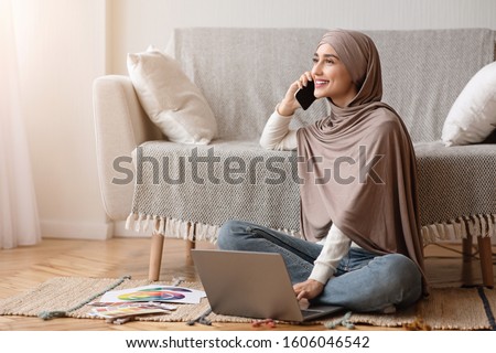 Photo of Work Opportunities For Muslim Women Concept. Smiling Arabic Girl In Hijab Talking On Cellphone And Using Laptop While Sitting On Floor At Home.