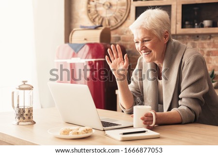 Elderly woman making video call on laptop in kitchen, waving at screen, chatting with children, free space