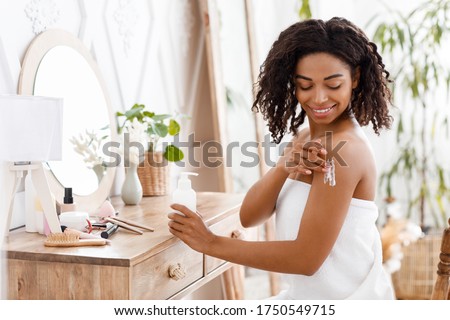 Skin Nutrition. Smiling Attractive Black Woman In Towel Applying Body Lotion, Pampering Herself After Bath, Sitting Wrapped In Towel At Dressing Table 商業照片 © 