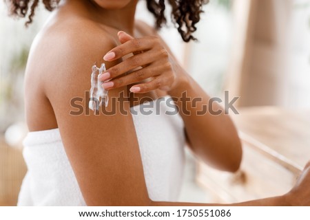 Skin Care Products Concept. Black woman applying moisturizing lotion on body after shower, standing wrapped in towel, cropped image