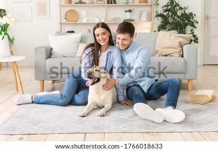 Happy Loving Family. Portrait of beautiful spouses patting dog sitting on the floor carpet in modern apartment