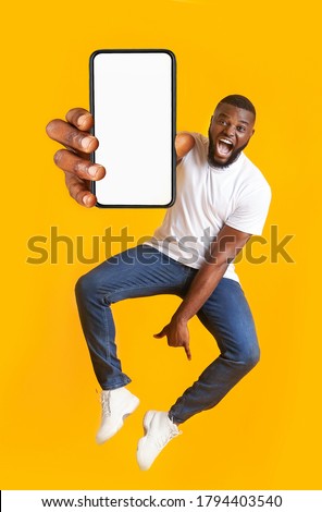New awesome mobile app. Closeup of smartphone with blank screen in jumping emotional black guy hand, yellow studio background