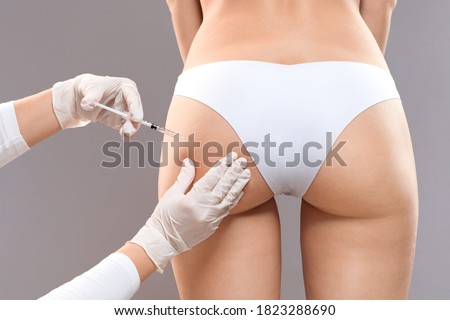 Sculptra butt lifting concept. Slim woman having hip injection at beauty salon, closeup. Plastic surgeon making injection at butt area for unrecognizable lady, grey studio background, back view