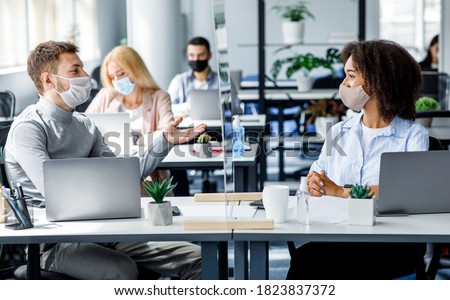 Communication and meeting in office after returning from covid-19 quarantine. Young guy and african american woman in protective mask talking through glass board at workplace with laptops in office