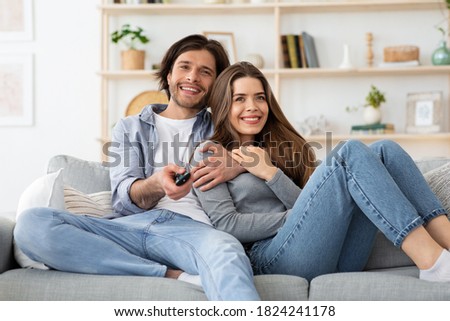Joyful young couple watching TV together at home, embracing and sitting on couch. Cheerful man and woman switching channels on TV with remote, seeking nice movie for weekend, empty space