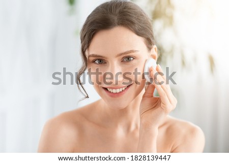 Smiling pretty young woman cleaning her face, using cotton pads and cleansing product, closeup. Young attractive lady using face toner and cotton pad, home interior, empty space, face care products