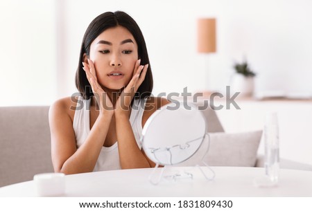 Facelift And Skin Care Concept. Portrait Of Young Asian Lady Massaging Her Face Sitting In Front Of The Mirror. Lady Applying Moisturizing Cream, Doing Facial Anti Aging Massage At Home