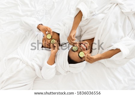 Beauty Spa Day. Cheerful Black Mom And Daughter In Bathrobes Lying With Cucumber Slices On Eyes, Doing Face Mask Treatment, Wearing Towel On Head, Having Fun Together At Home, Top View With Copy Space