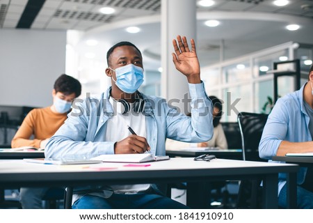 Education, Pandemic And Health Concept. African american male student raising hand for answer, wearing protective medical mask for protection from virus disease at high school lesson. New Normal
