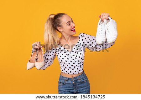 Making Decision. Portrait of smiling lady holding pair of high heels shoes and white sneakers in hands, can't choose between footwear and style, isolated over orange studio background Foto stock © 
