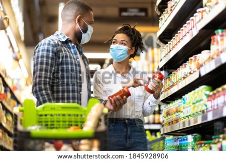 Grocery Shopping. Black Family Couple In Masks Buying Groceries In Supermarket Store Indoors. Buyers Standing With Shopping Cart In Aisles Choosing Food Products. Shop Safe During Covid-19 Pandemic