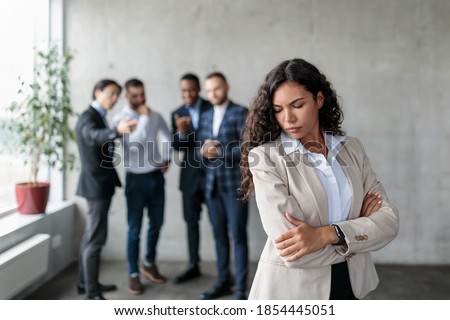 Workplace Sexism And Bullying. Unhappy Victimized Businesswoman Standing While Her Male Colleagues Whispering Behind Her Back Standing In Modern Office. Corporate Problems, Female Rights Concept