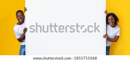 Cheerful african american couple standing by empty board for advertisement or text over yellow studio background. Smiling black man and woman standing next to blank placard for advert, panorama 商業照片 © 
