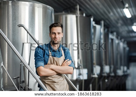 Work in modern brewery and favorite job. Confident attractive smiling guy brewer or owner in apron and controls equipment at factory with large boilers for brewing beer in plant interior, free space ストックフォト © 