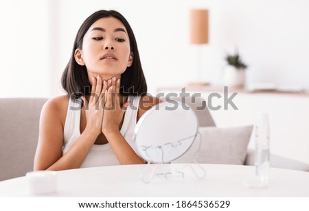 Facelift And Skin Care Concept. Portrait Of Beautiul Asian Woman Massaging Her Neck Sitting In Front Of The Mirror. Lady Applying Moisturizing Cream, Doing Anti Wrinkle Massage At Home