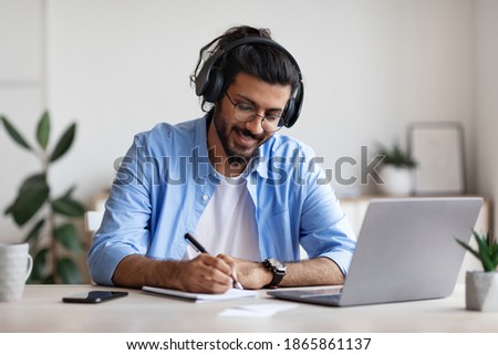Happy Western Guy Wearing Headphones Studying Online With Laptop At Home, Smiling Millennial Indian Guy Watching Webinar And Taking Notes To Notepad, Enjoying Distance Learning, Free Space