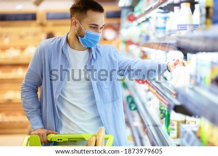 Consumerism And Consumption. Male customer in medical face mask choosing dairy products, taking bottle of milk or yoghurt from the fridge. Man standing with shopping trolley near freezer in mall