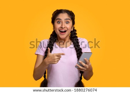 Yes, Great News. Portrait Of Excited Indian Woman Celebrating Win, Using Smartphone And Pointing At Gadget, Smiling Young Lady Having Success, Holding Cellphone, Yellow Studio Background