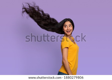 Portrait of smiling indian model woman posing with long and healthy dark blowing flying hair. Beautiful young lady looking back isolated over purple studio background, copy space. Haircare Advert